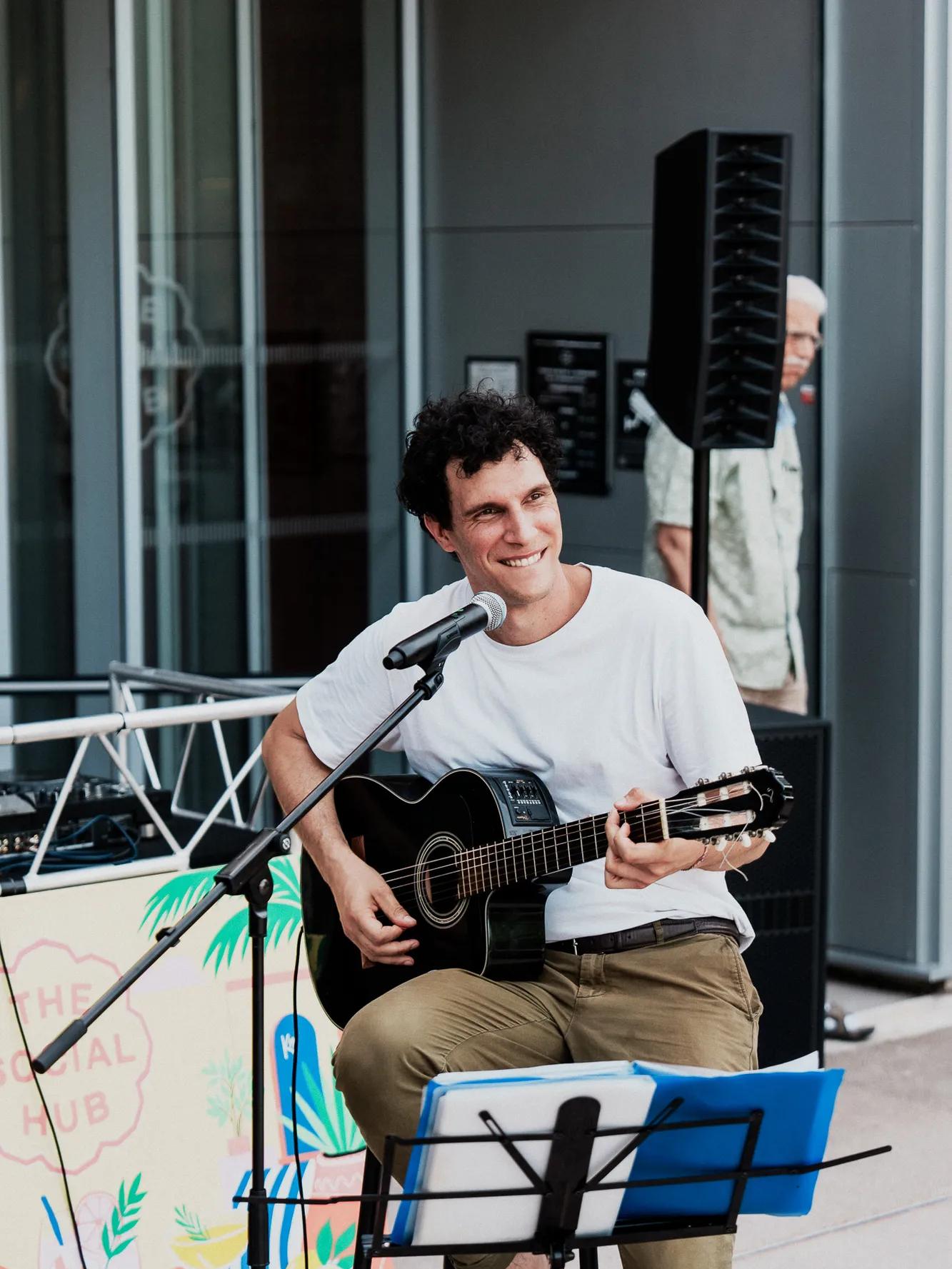 A smiling man in a white shirt plays a black acoustic guitar and sings into a microphone while seated outdoors. A music stand and a colorful sign are nearby.