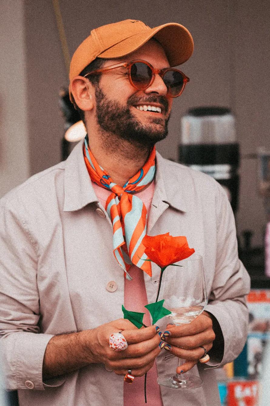 A smiling man wearing sunglasses, a cap, and a scarf holds a red paper rose while standing indoors, with blurred people and magazines in the background.