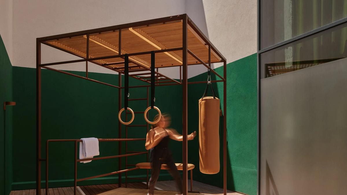 A person in activewear punches a hanging boxing bag in a modern gym area with gymnastics rings, a towel, and dumbbells on a wooden floor. The setting has green and white walls and a glass window.