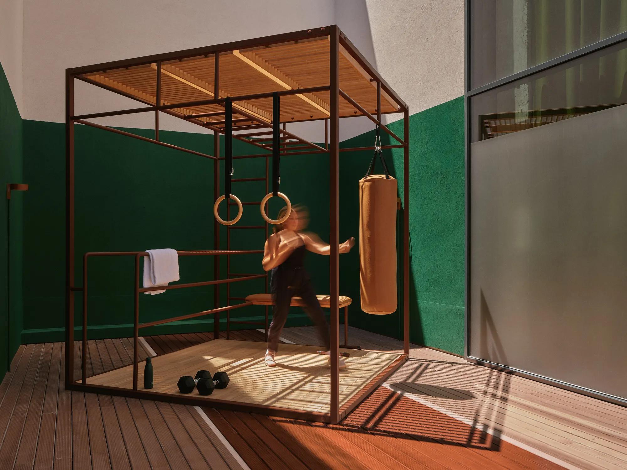 Una persona vestida con ropa deportiva golpea un saco de boxeo colgado en un gimnasio moderno, con anillas de gimnasia, una toalla y mancuernas sobre un suelo de madera. El entorno tiene paredes verdes y blancas y una ventana de cristal.