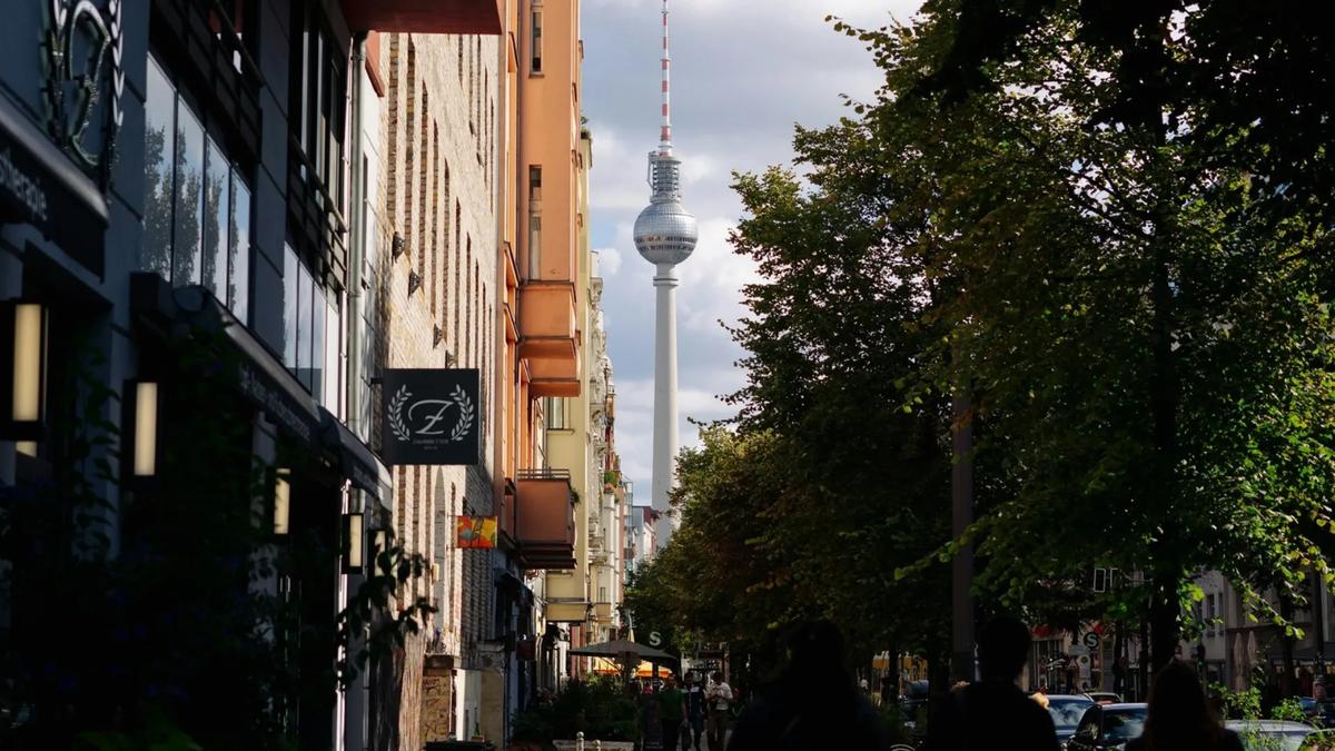 A city street lined with trees and buildings leads toward a distant television tower under a cloudy sky. The scene is lively with people walking along the sidewalk.