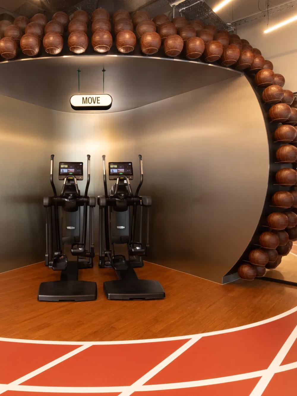 Two elliptical machines stand under an arched structure covered with basketballs, inside a modern gym with a running track design on the floor. A sign above reads MOVE. Glass-walled rooms are visible on both sides.