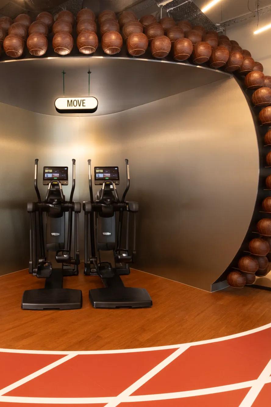 Deux vélos elliptiques sont installés sous une structure voûtée recouverte de ballons de basket, à l'intérieur d'une salle de sport moderne dont le sol est dessiné comme une piste de course. Au-dessus, un panneau porte l'inscription « MOVE ». Des salles aux parois vitrées sont visibles de part et d'autre.
