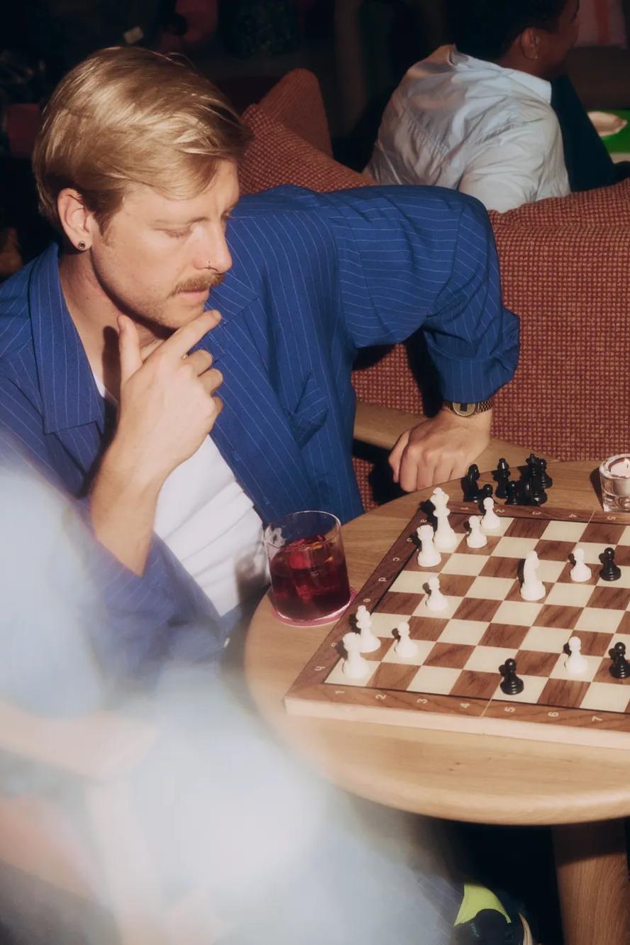 Two people sit at a small table, playing chess. One person ponders a move, while the other sips a cocktail. Both appear relaxed in a cozy, softly lit setting with others in the background.