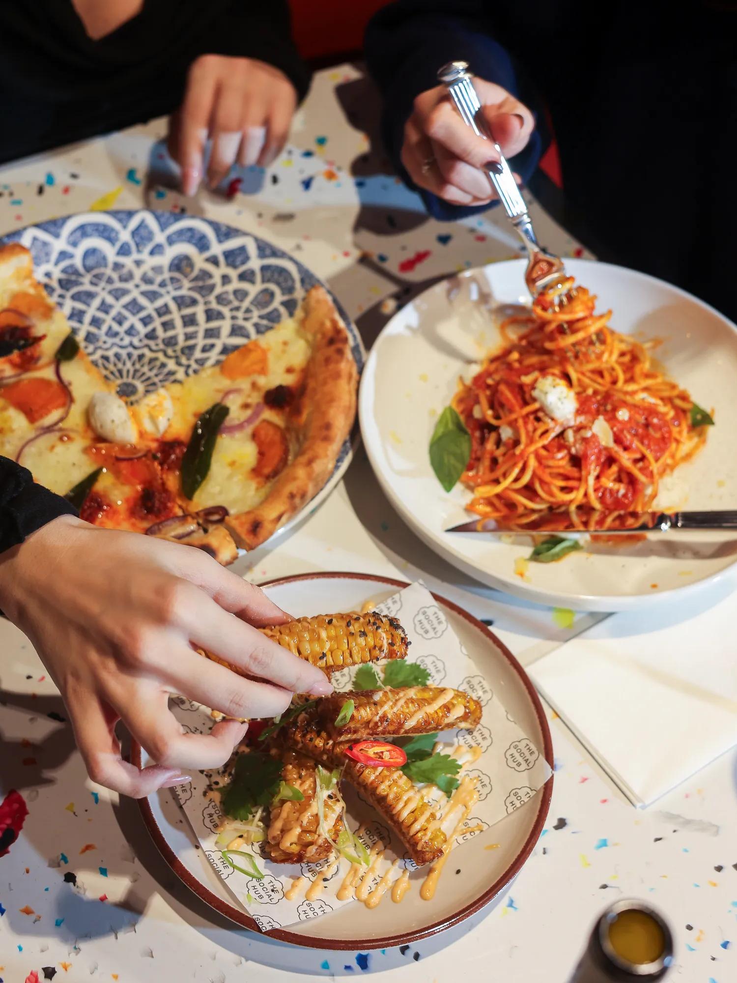 Two people share a meal with pizza, spaghetti, and fried food on a confetti-covered table. One person twirls pasta with a fork, while another reaches for a piece of fried food.