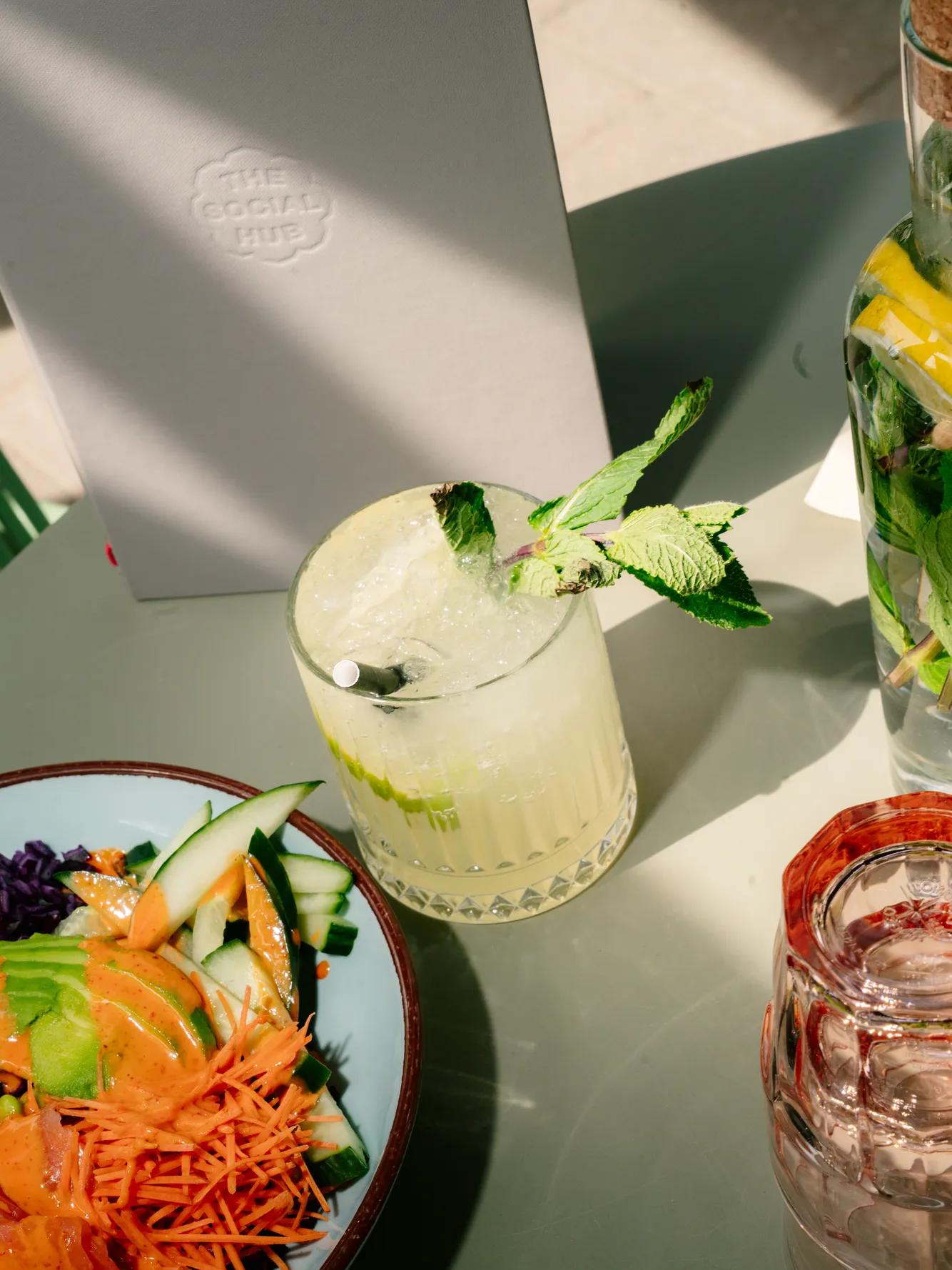 A glass of lemonade with mint, a bowl of salad with carrots and cucumber, a jar of lemon water, and a menu on a sunlit table.