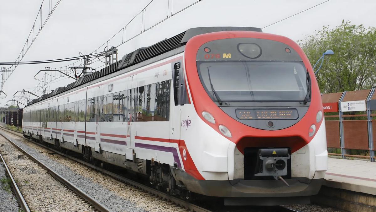 A modern white and red passenger train with renfe branding is stopped at an outdoor railway station, with overhead power lines and trees in the background.