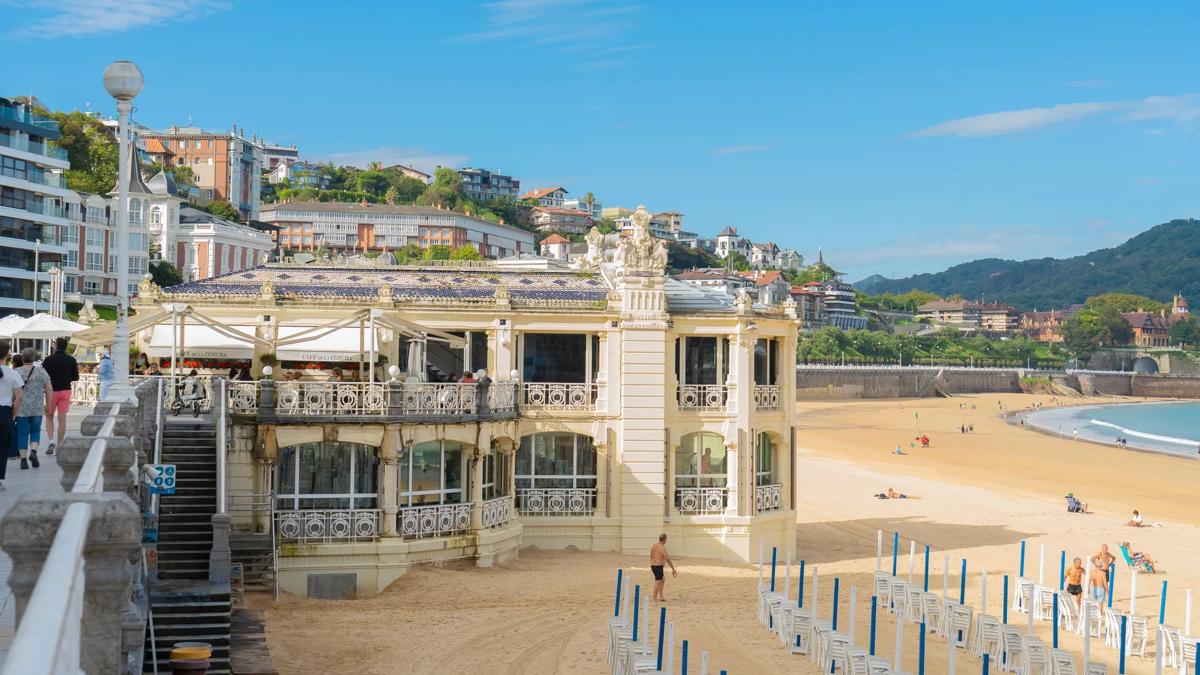 Elegant seaside building with decorative railings sits on a sandy beach. Blue sky, some clouds, and sunbathers are visible. Hills and distant houses rise in the background, and rows of closed umbrellas line the beach.