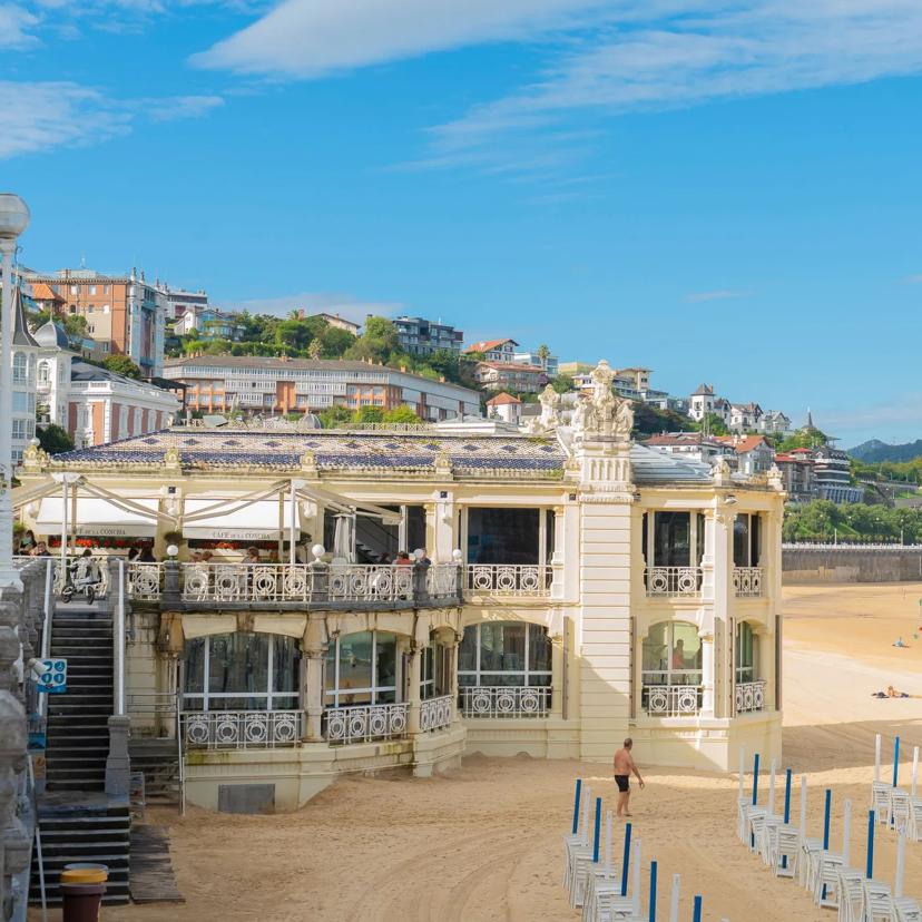 Elegant seaside building with decorative railings sits on a sandy beach. Blue sky, some clouds, and sunbathers are visible. Hills and distant houses rise in the background, and rows of closed umbrellas line the beach.