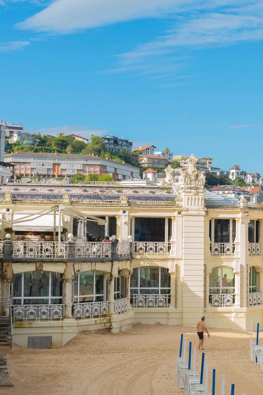 Ein elegantes Gebäude am Meer mit verzierten Geländern steht an einem Sandstrand. Zu sehen sind der blaue Himmel, einige Wolken und Sonnenbadende. Im Hintergrund erheben sich Hügel und Häuser in der Ferne, und am Strand reihen sich geschlossene Sonnenschirme aneinander.