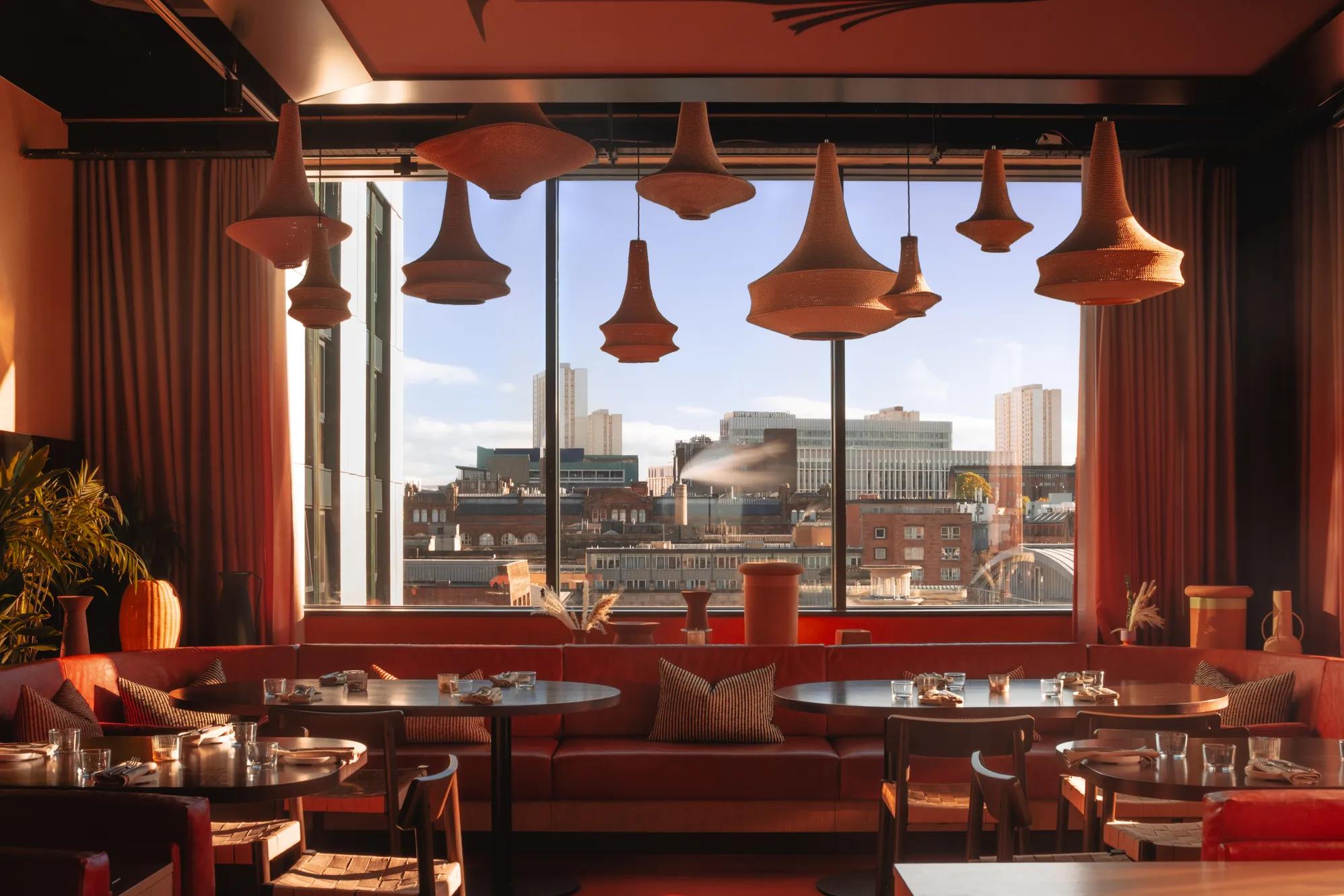 A stylish restaurant interior with warm lighting, woven hanging lamps, red seating, set tables, and large windows offering a city skyline view with modern buildings in the background.