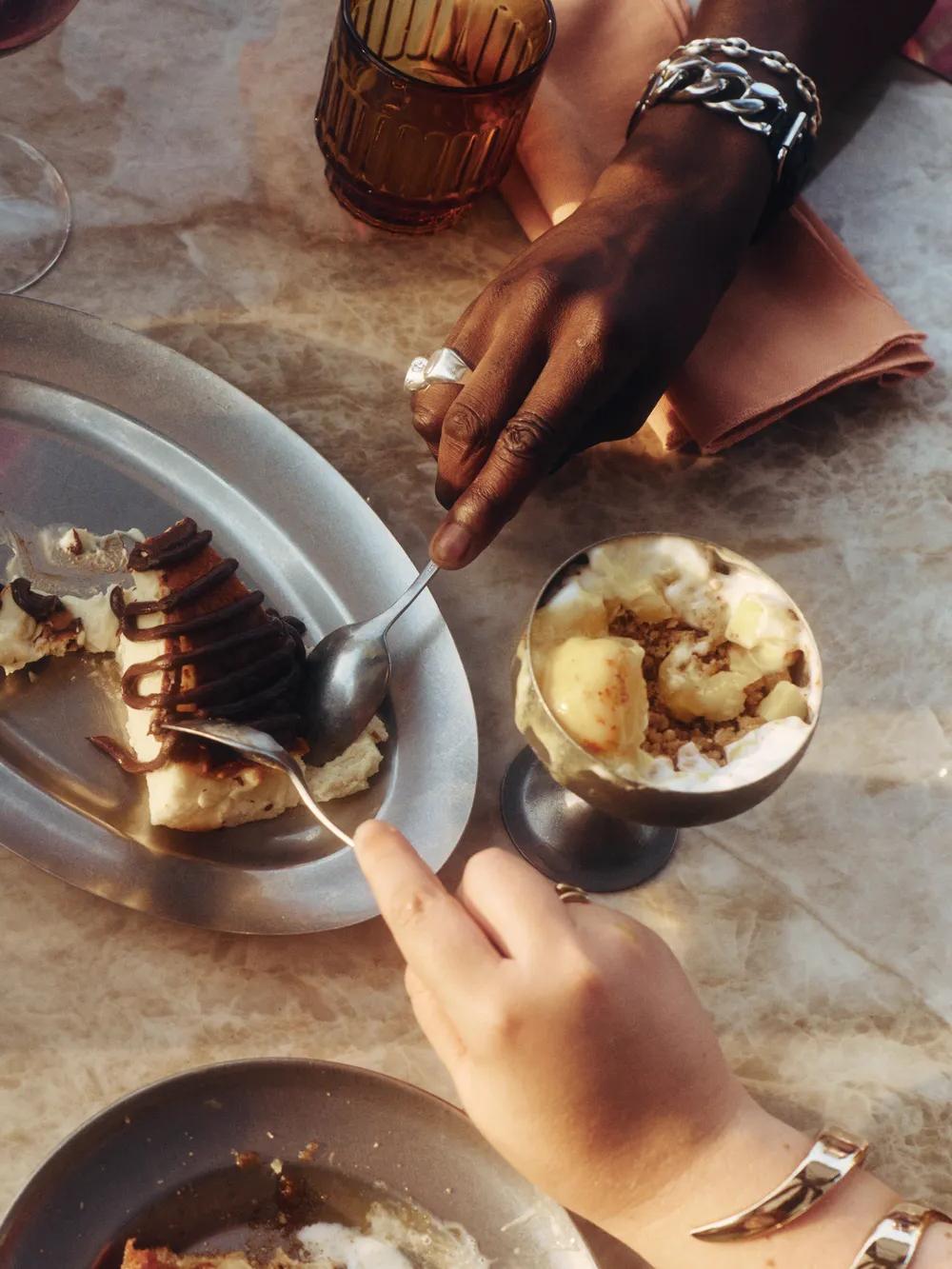 Three people share a dessert on a marble table, using forks to eat from a metal plate. There are drinks and a bowl of ice cream nearby. The scene appears warm and inviting.