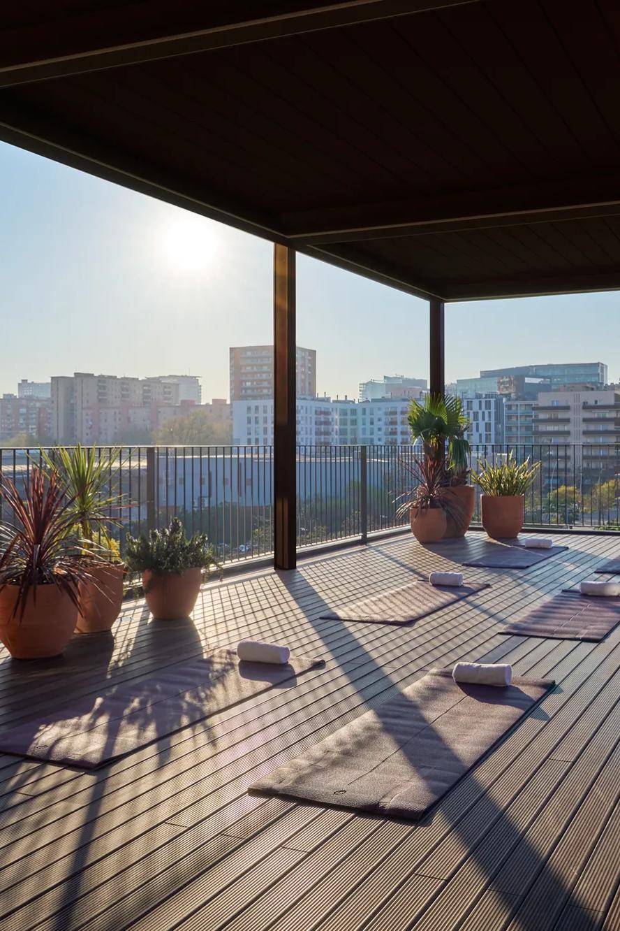 Yoga mats and blocks are arranged on a covered rooftop deck with potted plants, overlooking a cityscape in the morning sunlight.