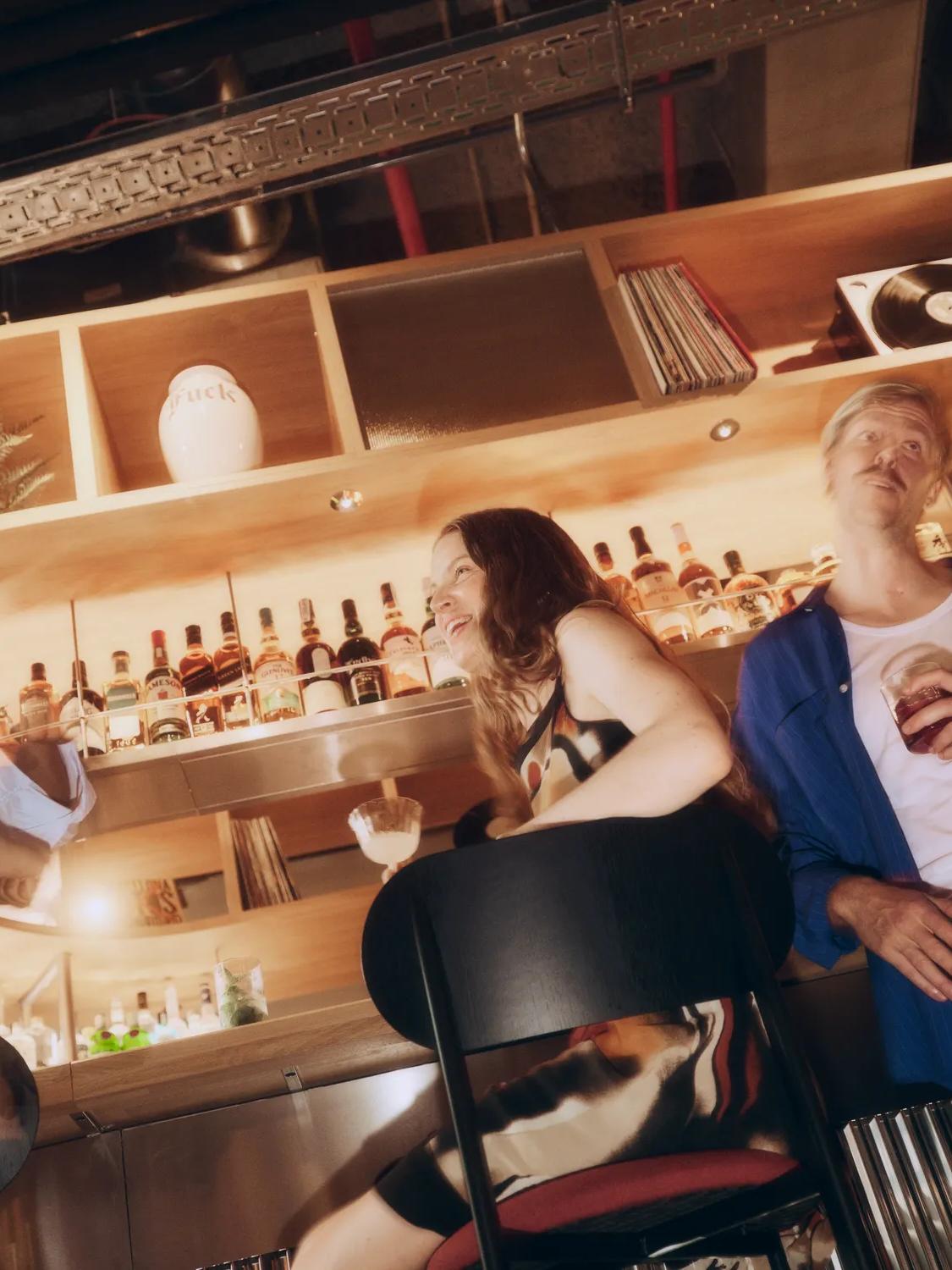 Four people socialize at a stylish bar, drinks in hand. Bottles, plants, and a record player decorate the wooden shelves behind them. The lighting is warm and the scene appears lively and casual.