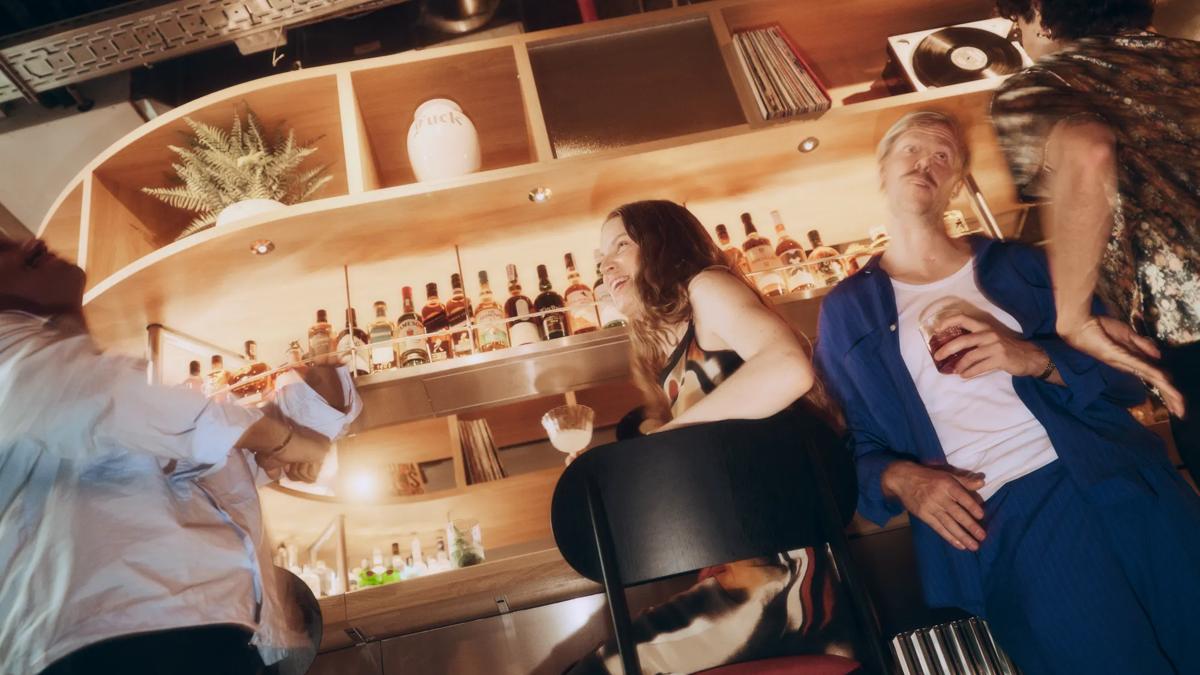Four people socialize at a stylish bar, drinks in hand. Bottles, plants, and a record player decorate the wooden shelves behind them. The lighting is warm and the scene appears lively and casual.