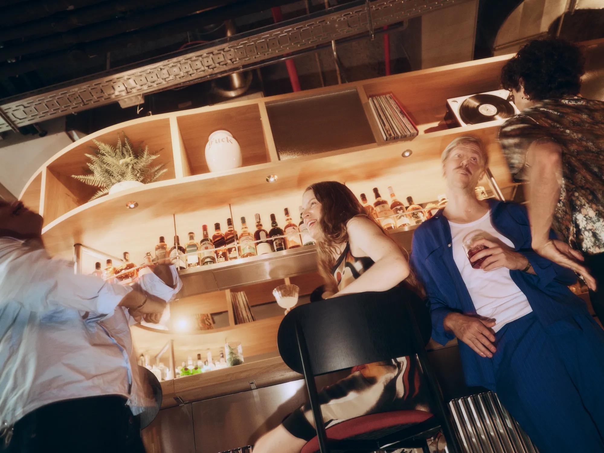 Cuatro personas charlan en un bar con mucho estilo, con una copa en la mano. Detrás de ellos, las estanterías de madera están decoradas con botellas, plantas y un tocadiscos. La iluminación es cálida y la escena transmite un ambiente animado y desenfadado.