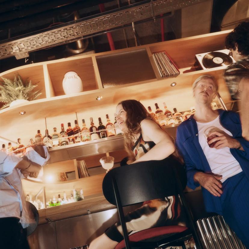 Cuatro personas charlan en un bar con mucho estilo, con una copa en la mano. Detrás de ellos, las estanterías de madera están decoradas con botellas, plantas y un tocadiscos. La iluminación es cálida y la escena transmite un ambiente animado y desenfadado.