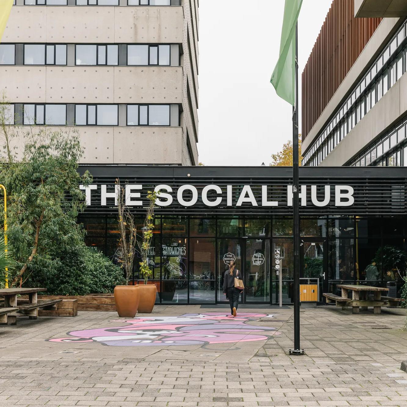Entrance of The Social Hub, with benches, greenery, tall flagpoles, and a colorful painted design on the pavement in front of a modern building. A person is walking toward the glass doors.