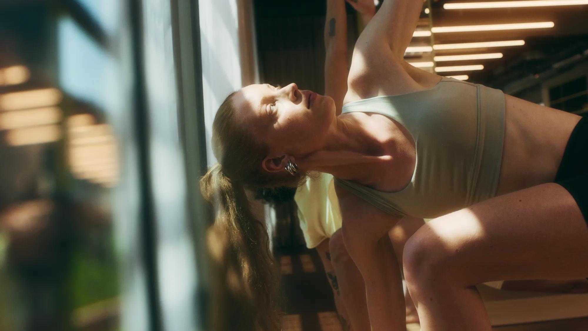 Woman in athletic wear doing a side stretch yoga pose indoors, with sunlight streaming in through large windows. Other participants are partially visible in the background.