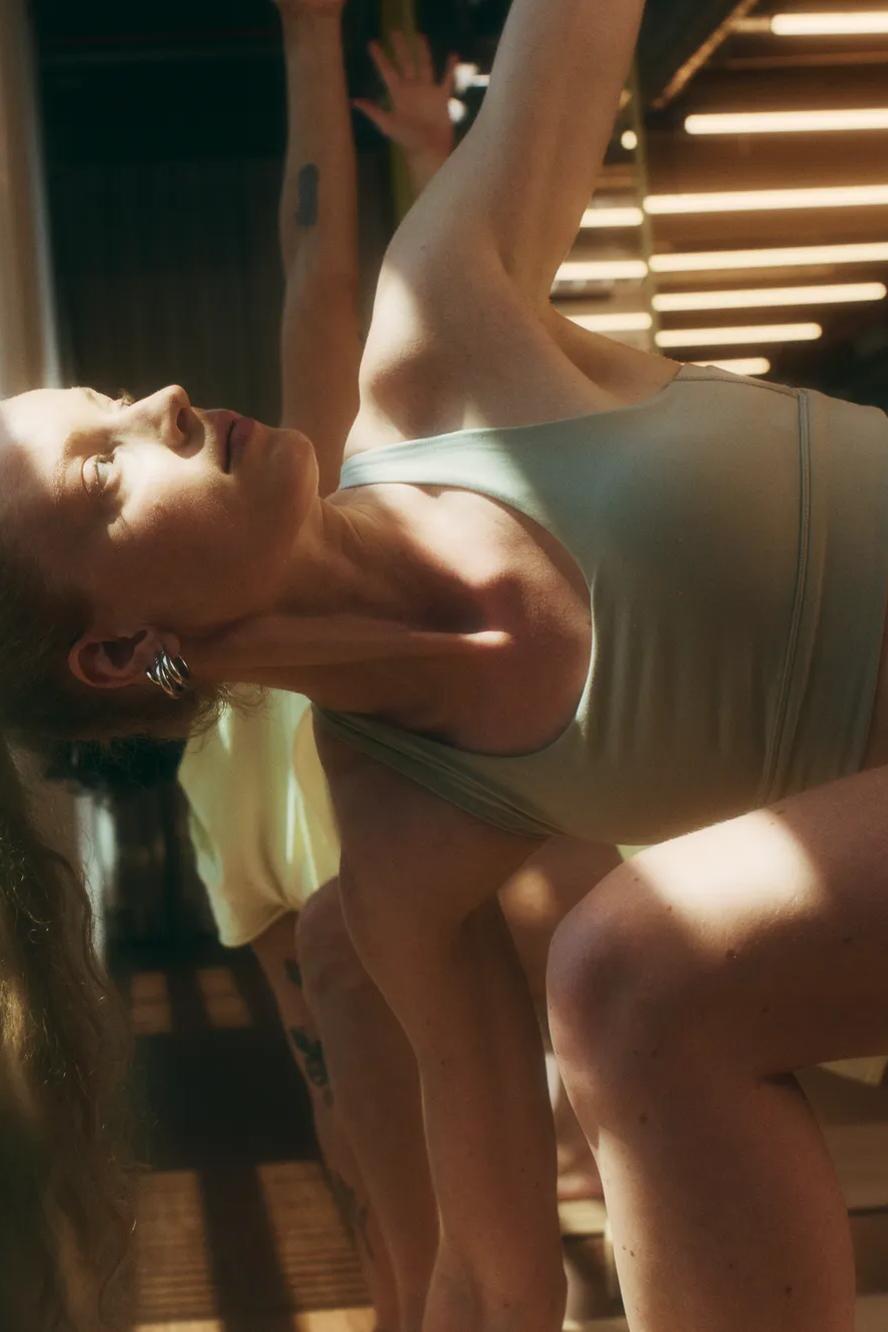 Woman in athletic wear doing a side stretch yoga pose indoors, with sunlight streaming in through large windows. Other participants are partially visible in the background.