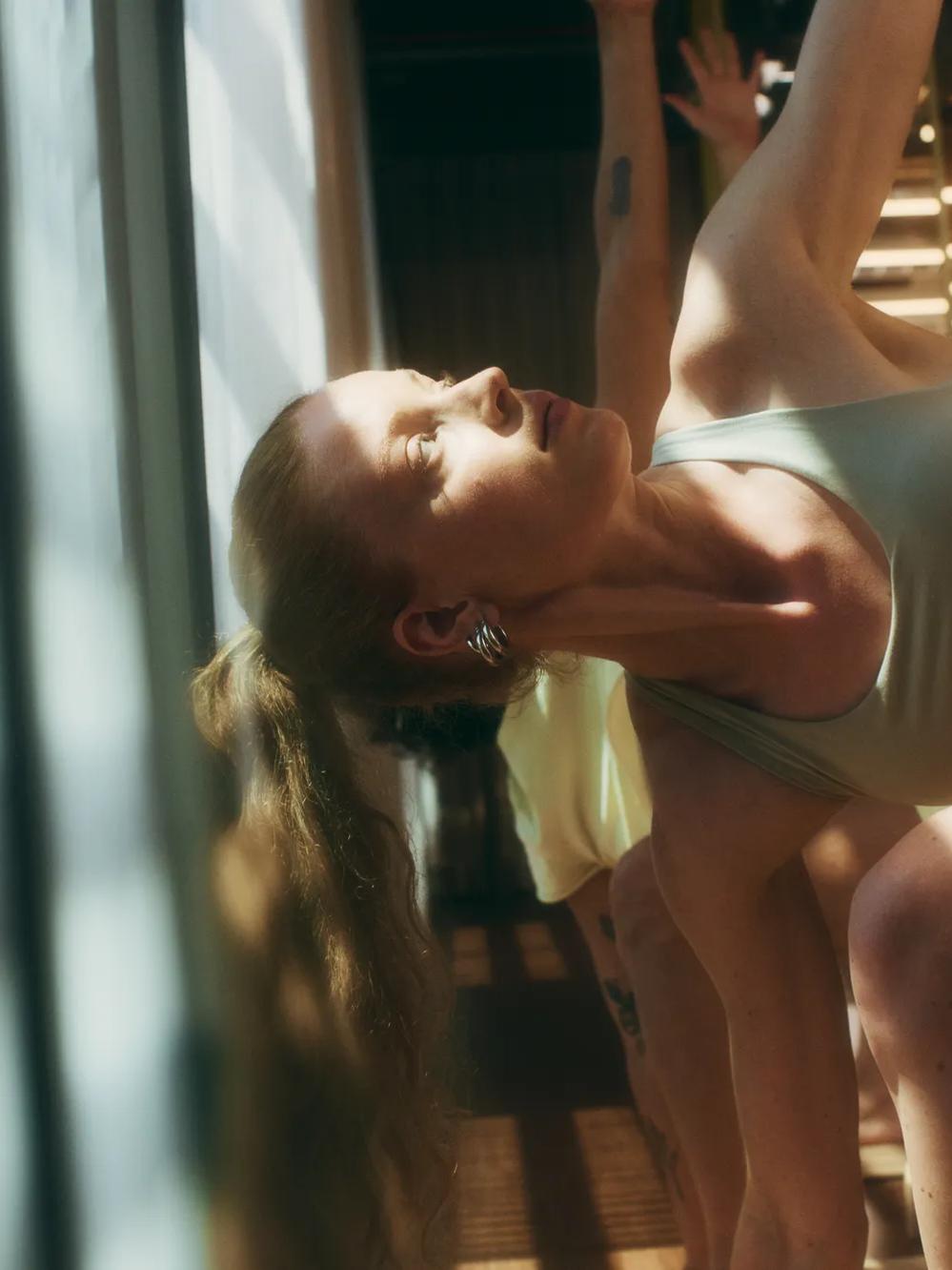 Woman in athletic wear doing a side stretch yoga pose indoors, with sunlight streaming in through large windows. Other participants are partially visible in the background.