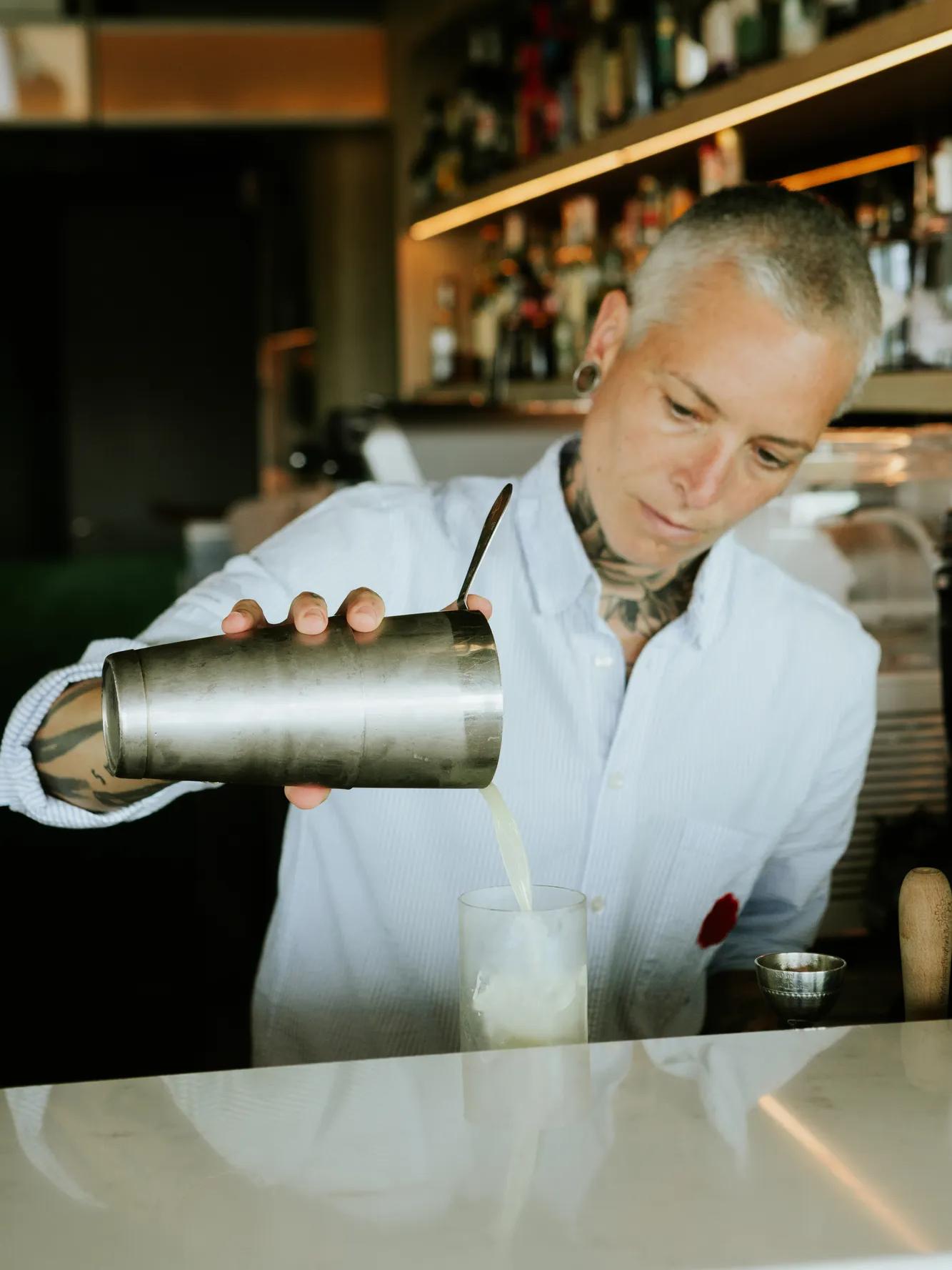 A person with short hair and tattoos pours a drink from a metal shaker into a glass at a bar, with bottles visible in the background.