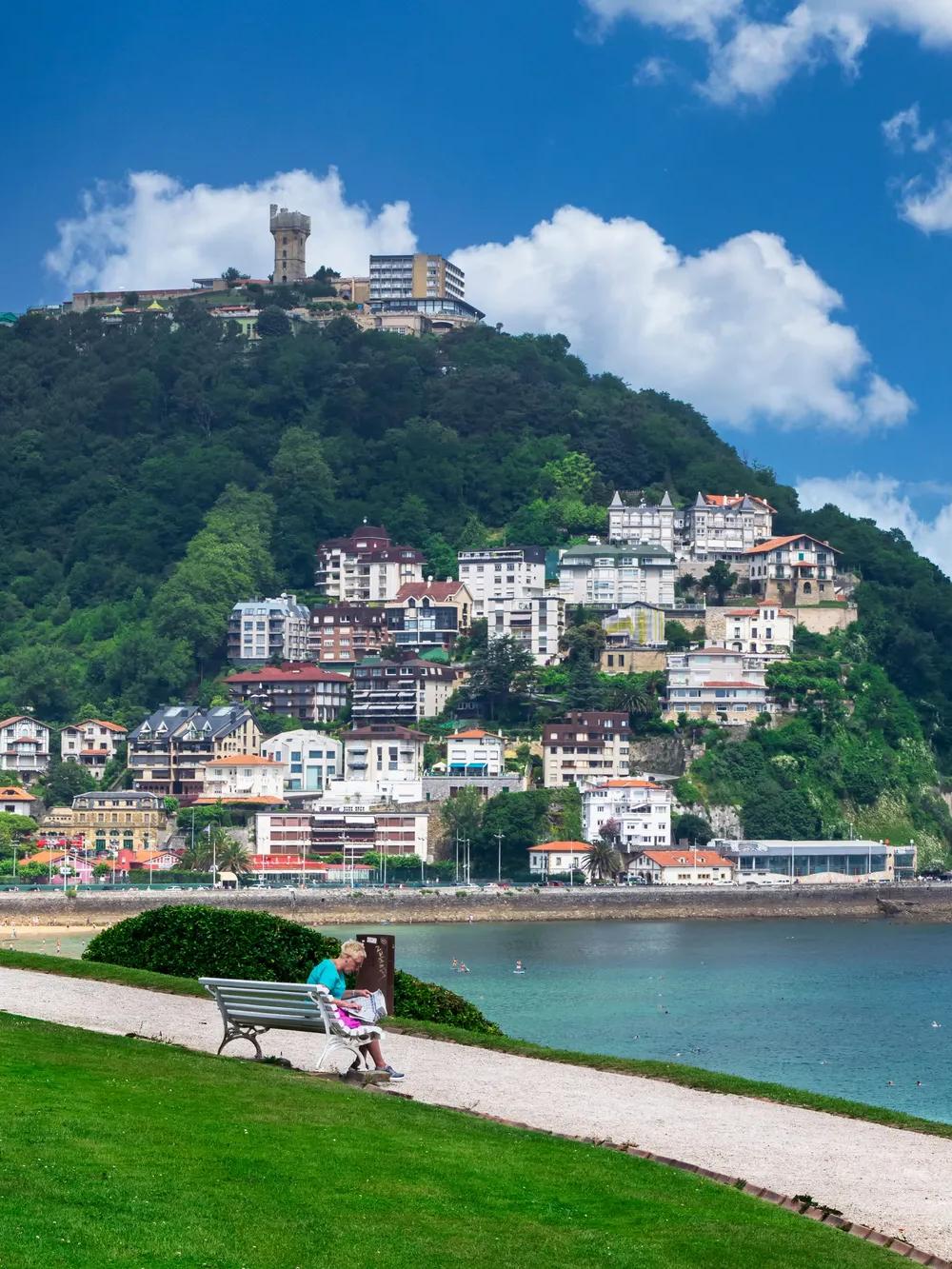 A person sits on a bench facing the ocean, with colorful buildings on a green hillside and a tower on top. The sky is blue with scattered clouds, and the scene is peaceful and scenic.