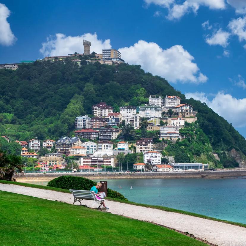 Eine Person sitzt auf einer Bank mit Blick auf das Meer; im Hintergrund sind bunte Gebäude an einem grünen Hang zu sehen, auf dessen Spitze ein Turm steht. Der Himmel ist blau und mit vereinzelten Wolken übersät, und die Szene wirkt friedlich und malerisch.