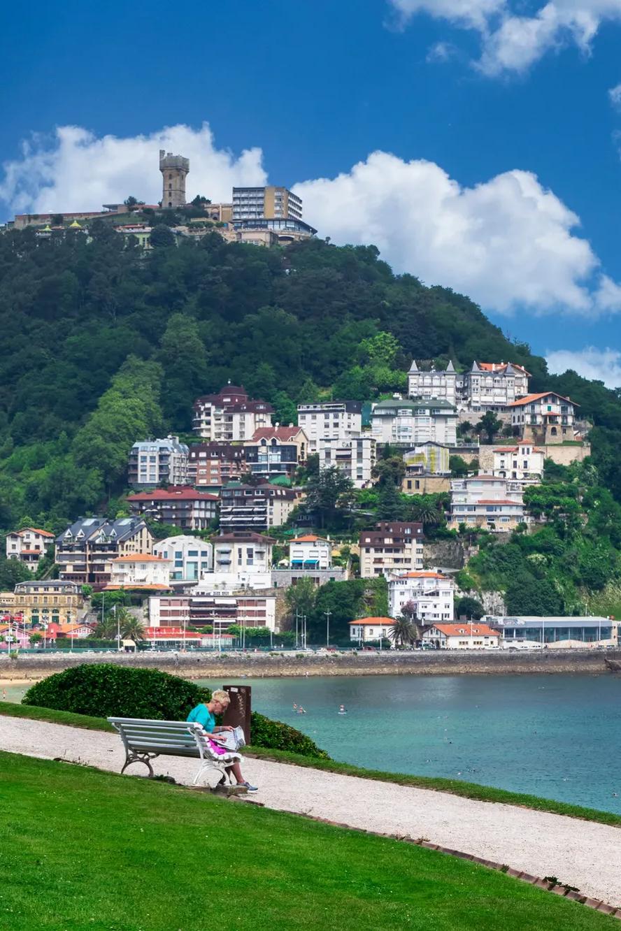 A person sits on a bench facing the ocean, with colorful buildings on a green hillside and a tower on top. The sky is blue with scattered clouds, and the scene is peaceful and scenic.