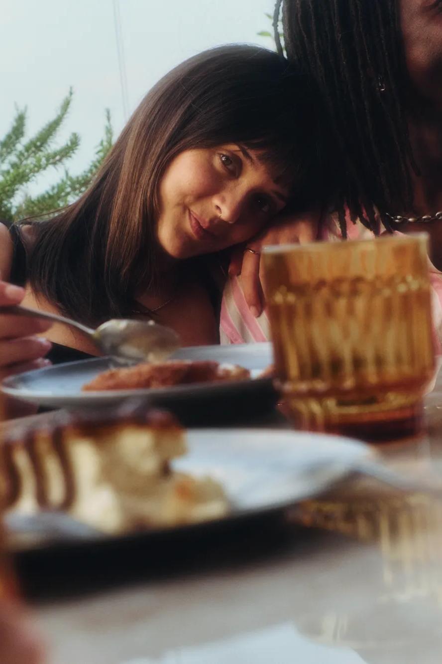 A woman rests her head on a mans shoulder at an outdoor table set with desserts and drinks, both smiling and enjoying a relaxed moment together.