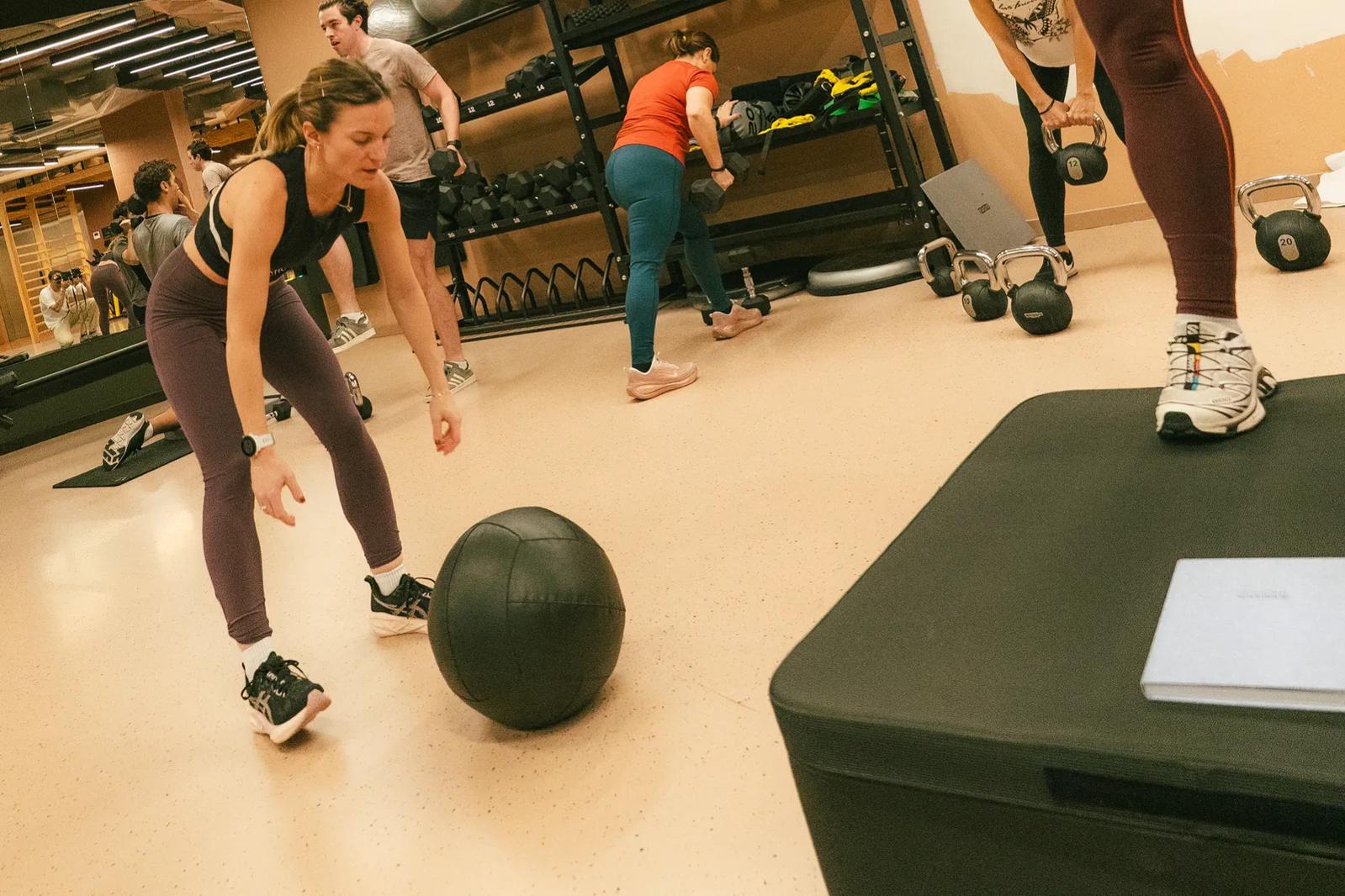 People exercise in a gym; one woman steps on a platform and smiles, another bends to pick up a medicine ball, while others lift weights in the background. The room has gym equipment and bright overhead lights.