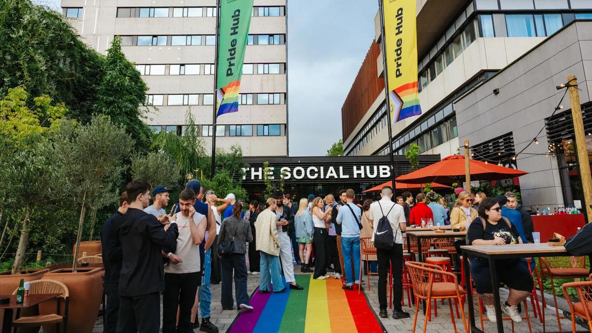 People gather outside The Social Hub, standing on a rainbow-colored carpet flanked by plants, with tall buildings and colorful The Social Hub banners in the background.