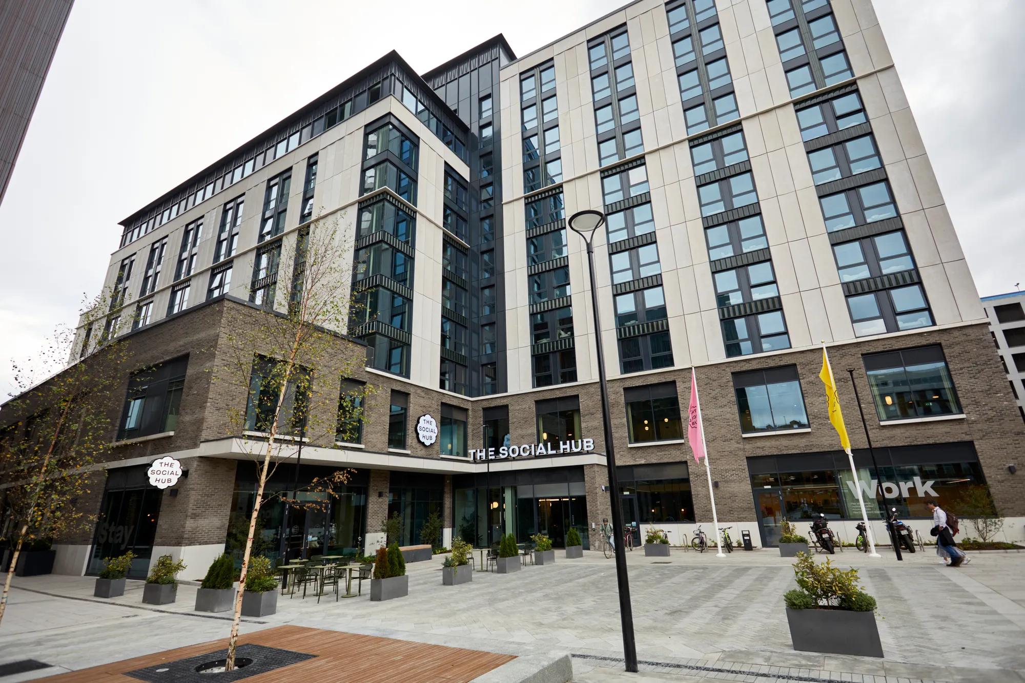 Modern multi-story building with large windows, sign reading The Social Hub, outdoor seating, trees, and people walking in a spacious courtyard with pavers.