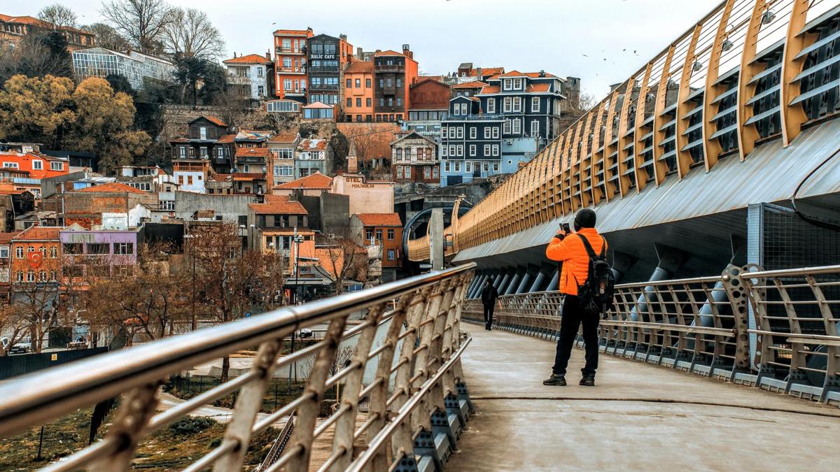 A person in an orange jacket takes a photo on a bridge with metal railings, overlooking colorful hillside buildings in a European city.