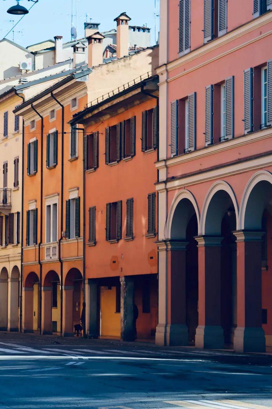 In una giornata di sole, edifici dai colori vivaci con portici ad arco e finestre con le persiane si affacciano su una strada tranquilla; si intravedono alcune auto parcheggiate e le ombre si allungano sulla strada.