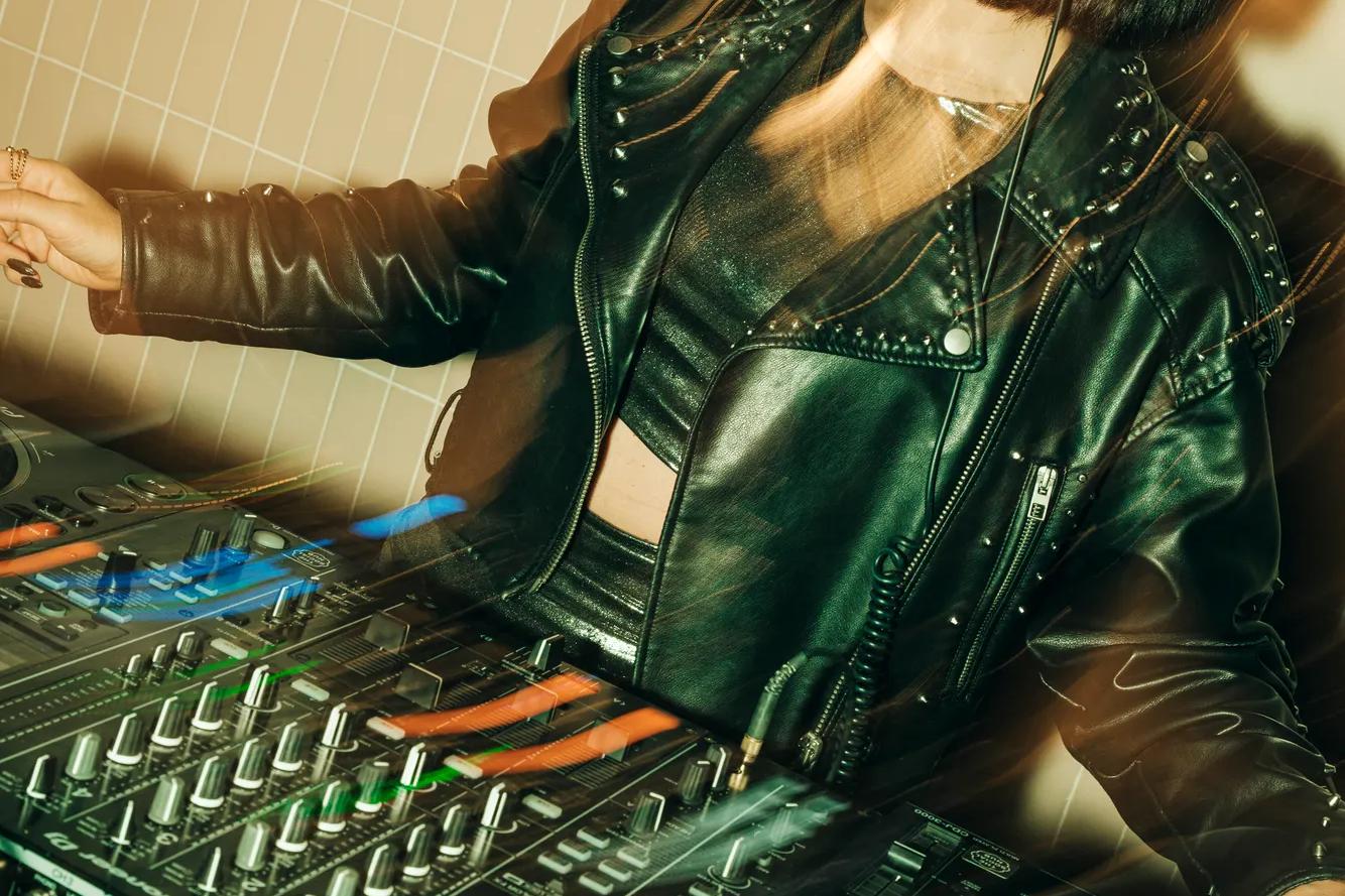 A woman in a black leather jacket and headphones is DJing at a mixer. The lighting creates a motion blur, giving the scene an energetic and dynamic feel.