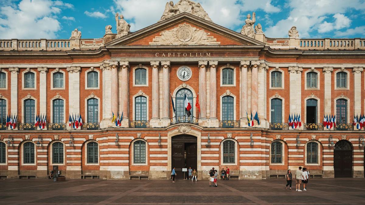 The Capitole de Toulouse, a grand historic building with a red and white brick facade, under a blue sky with pedestrians walking in the plaza in front.