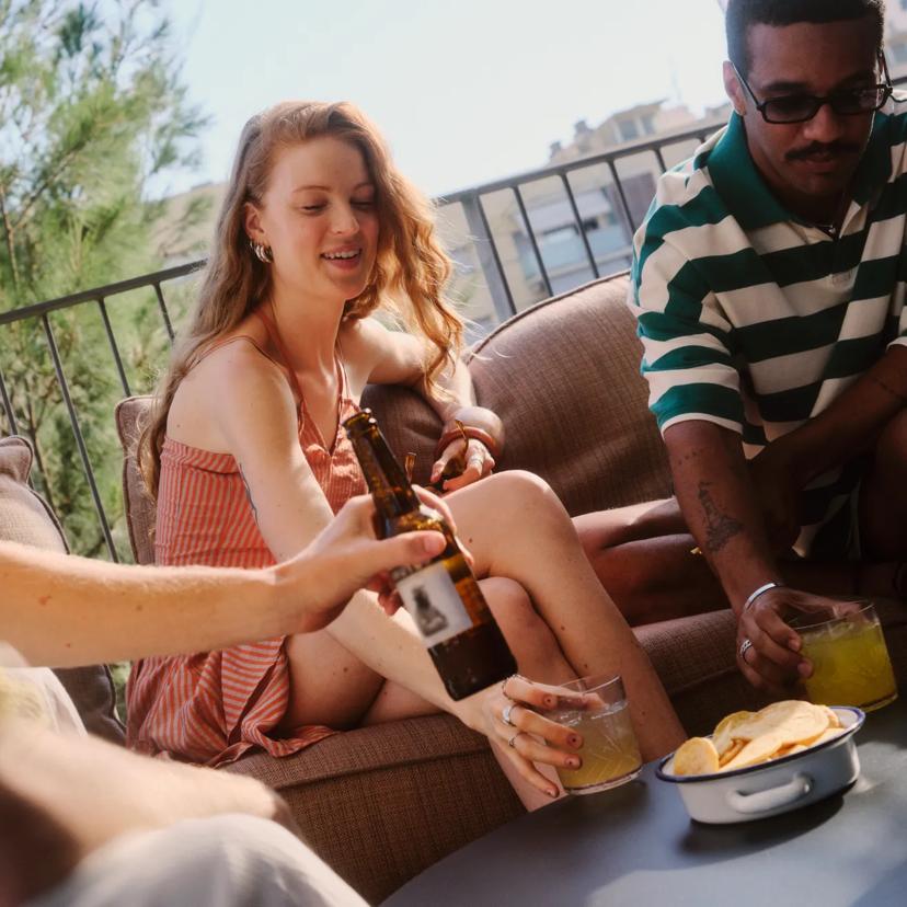 Three people relax on an outdoor patio couch, sharing drinks and snacks. One person pours beer, while another holds a glass. A bowl of chips is on the table, and sunlight filters through the trees.