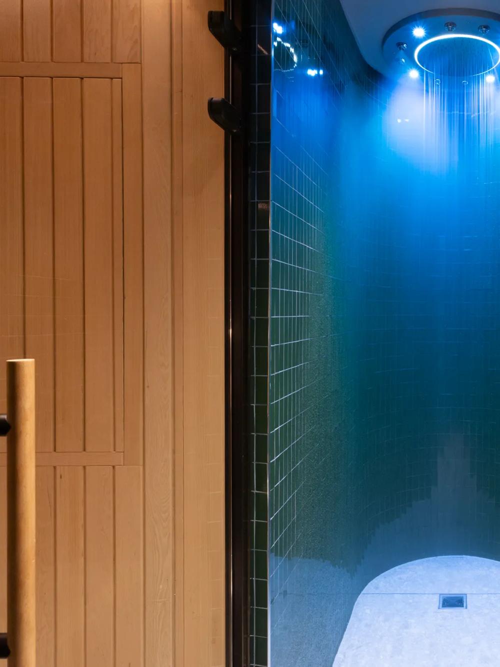 A modern spa shower with dark green tiles and a blue-lit rainfall showerhead sits between a wooden sauna door on the left and a light-colored relaxation area with a wooden bench on the right.