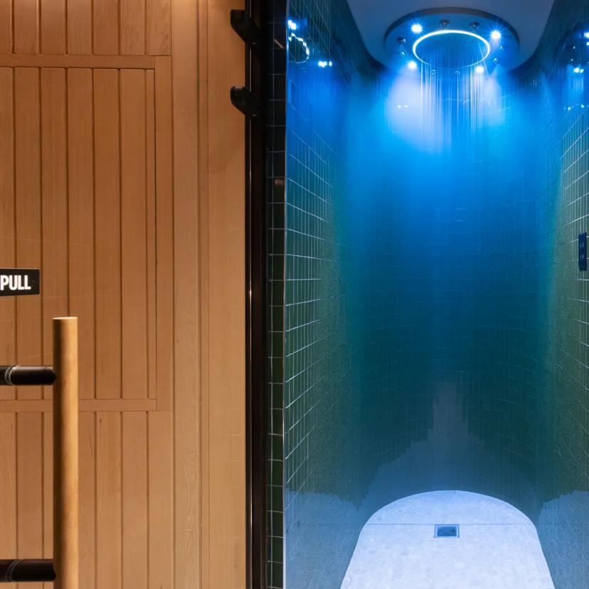 Eine moderne Wellnessdusche mit dunkelgrünen Fliesen und einem blau beleuchteten Regenduschkopf befindet sich zwischen einer Saunatür aus Holz auf der linken Seite und einem hellen Entspannungsbereich mit einer Holzbank auf der rechten Seite.