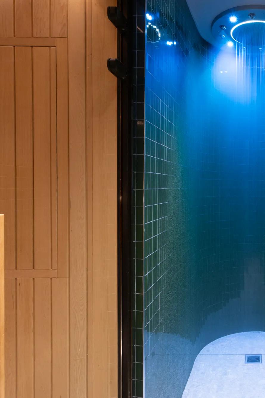 Una moderna doccia spa con piastrelle verde scuro e un soffione a pioggia illuminato di blu si trova tra una porta in legno che dà sulla sauna, a sinistra, e una zona relax dai toni chiari con una panca in legno, a destra.