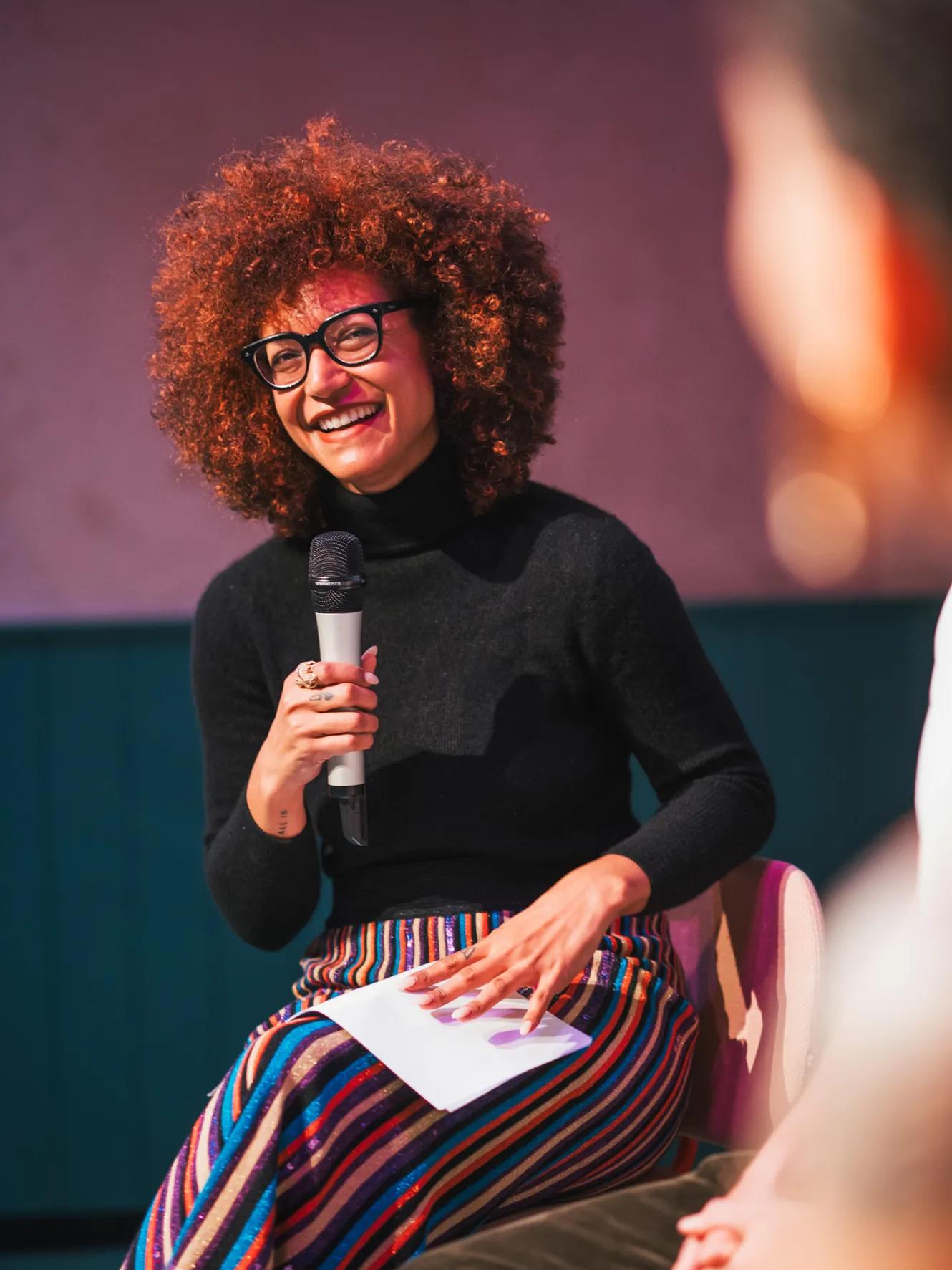 A smiling person with curly hair and glasses holds a microphone and paper, seated indoors. They wear a black turtleneck and colorful striped pants, appearing to engage in a lively discussion or interview.