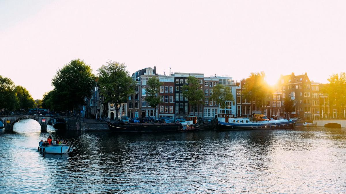 A small boat travels along an Amsterdam canal lined with historic buildings and trees at sunset.
