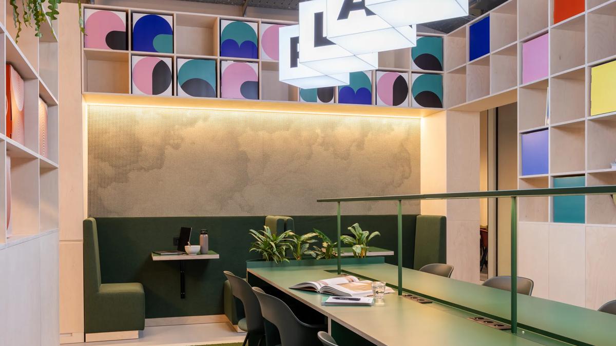 Modern workspace with green tables, matching chairs, booth seating, potted plants, and colorful geometric wall storage. Overhead, illuminated letter blocks hang from the ceiling. Open books and stationery are on the table.