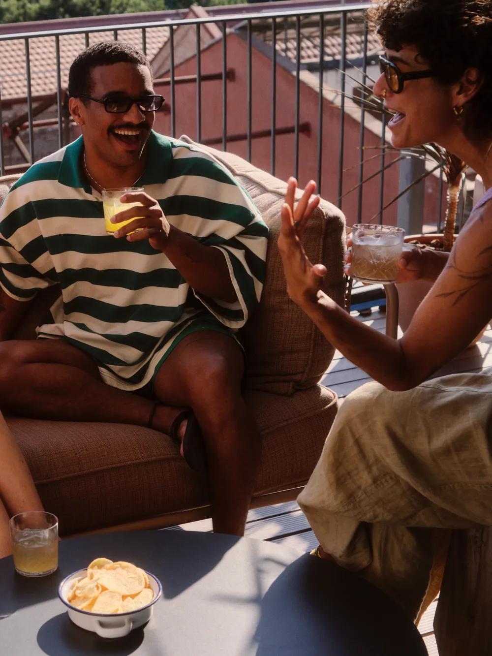 Four friends sit on an outdoor patio sofa, laughing and chatting while holding drinks. There is a bowl of chips on the table in front of them. Its a sunny day and they look relaxed and happy.