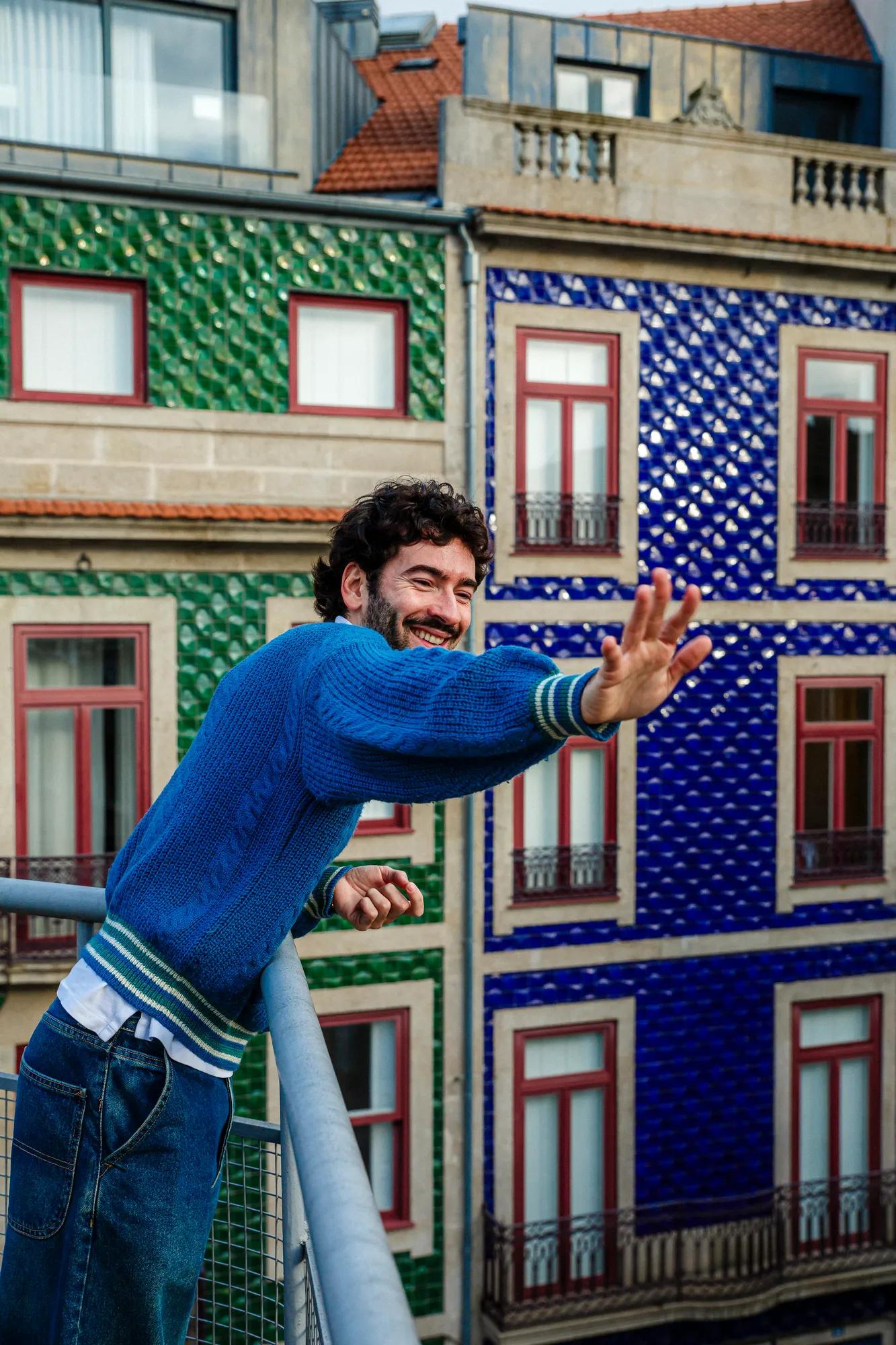 A man in a blue sweater smiles and waves from a balcony, with colorful green and blue tiled buildings in the background.