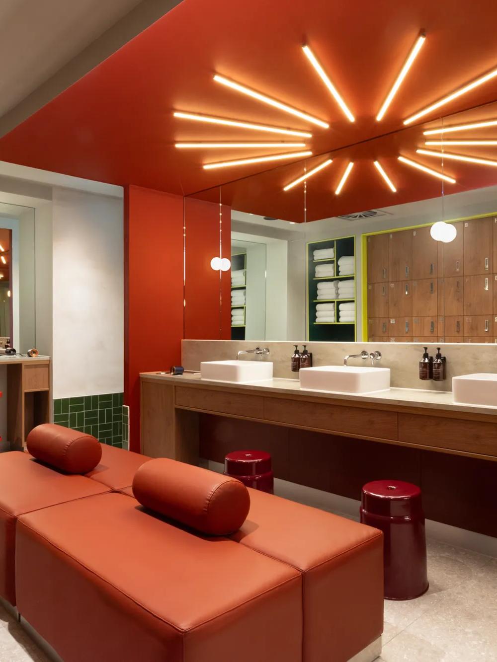 Modern restroom with red decor, starburst ceiling lights, three sinks, mirrors, red leather bench, stools, and wooden lockers along the back wall. Contemporary and stylish interior design.
