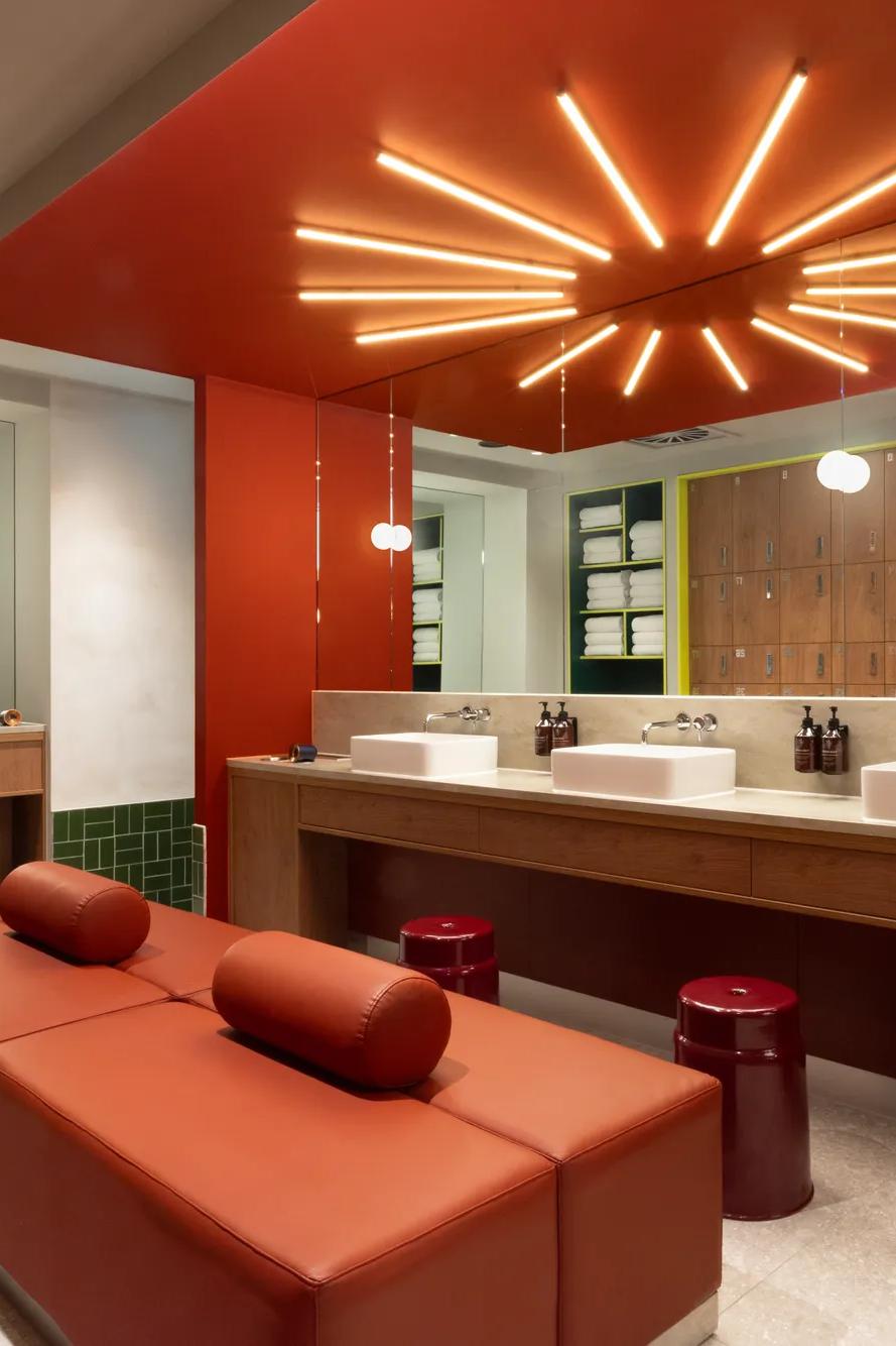 Modern restroom with red decor, starburst ceiling lights, three sinks, mirrors, red leather bench, stools, and wooden lockers along the back wall. Contemporary and stylish interior design.