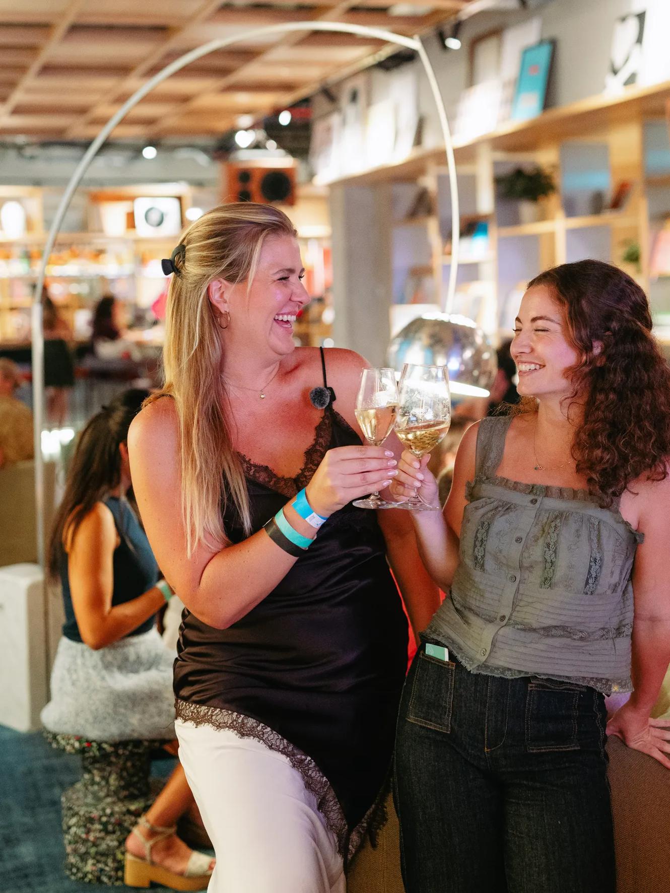 Two women smiling and toasting with glasses of white wine in a lively, modern indoor setting with shelves and people in the background.