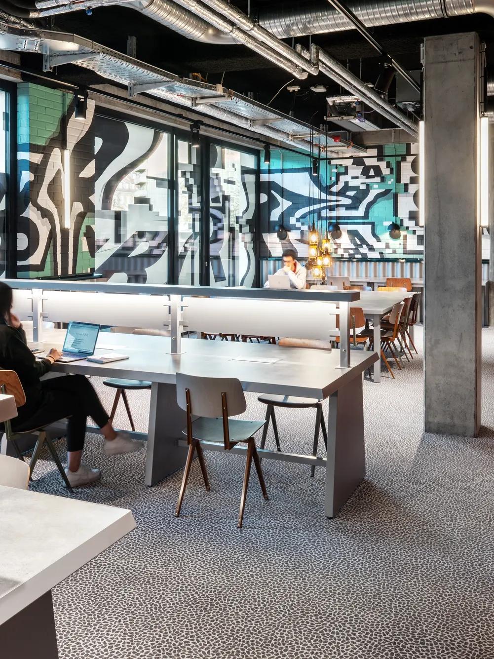 Modern office space with large windows, several people working at desks, industrial-style decor, visible ceiling pipes, and abstract black-and-white wall art in the background.